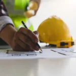 A person draws on architectural plans with a pencil, navigating building permits; a yellow hard hat and reflective vest are visible in the background.